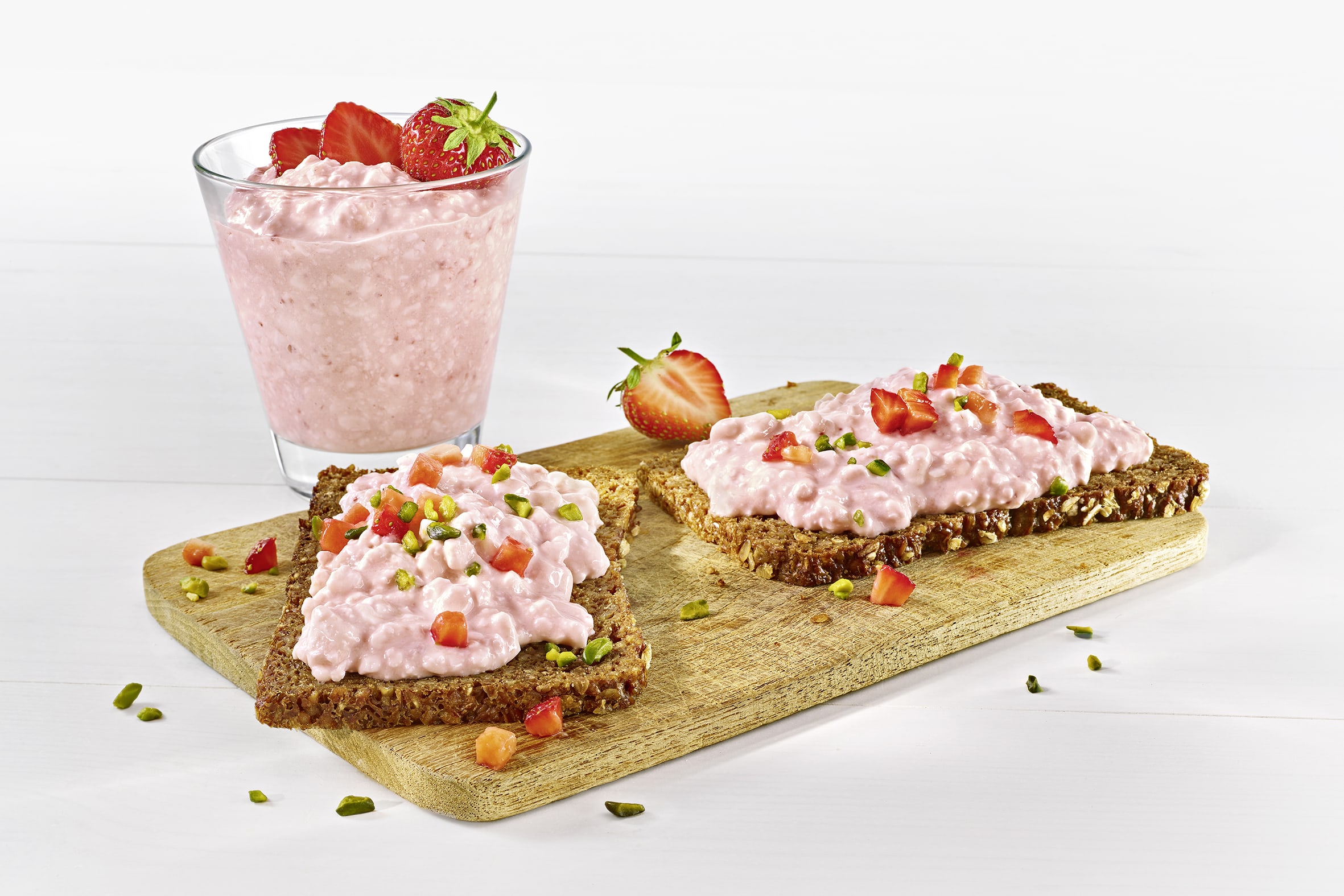 Zwei Scheiben Vollkornbrot mit körnigem Frischkäse und Erdbeersstücken auf einem Holzbrett. Dahinter steht ein Glase mit Körnigem Frischkäse und Erbeer-Topping. Zwei Scheiben Vollkornbrot mit körnigem Frischkäse und Erdbeersstücken auf einem Holzbrett. Dahinter steht ein Glase mit Körnigem Frischkäse und Erbeer-Topping.