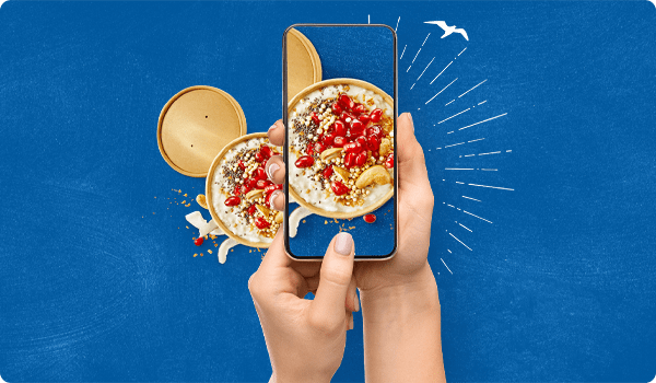 Zwei Hände, die ein Smartphone halten, auf dem ein To-Go-Becher mit Porridge, Granatapfelkernen, Chiasamen und Cashews zu sehen ist.