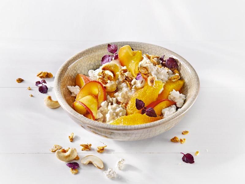 MILRAM Körniger Frischkäse in einer Bowl, garniert mit Orangenfilets, gedünsteten Apfelspalten, Granola, Cashewkernen und Shisokresse