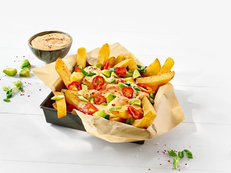 Pommes in einem eckigen Schälchen getoppt mit einer Käse-Chili-Sauce, Avocadowürfeln, Pfefferschoten und Frühlingszwiebeln. Daneben ein Schälchen mit Käsesauce.