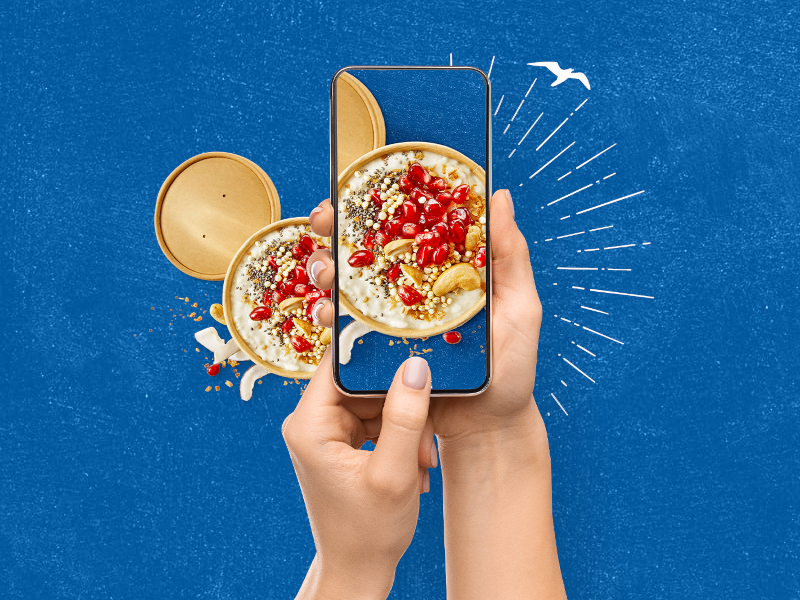 Zwei Hände halten ein Smartphone und fotografieren einen To-Go-Becher gefüllt mit MILRAM Porridge, Cranberrys, Cashewkernen und Chiasamen