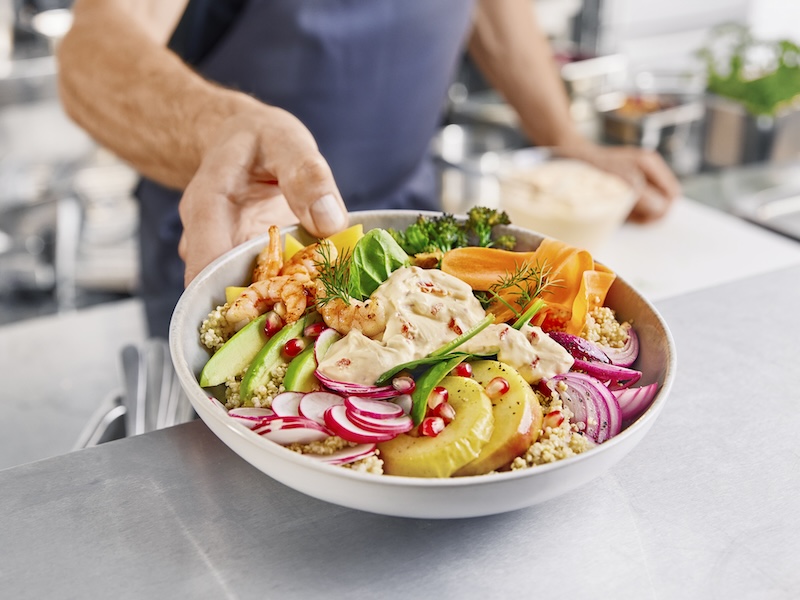 Schale gefüllt mit Quinoa, Avocado-Scheiben, gegrillten Apfelscheiben, Zwiebelringen, blanchiertem Bimi, gebratenen Garnelen, Granatapfelkernen und MILRAM PaprikaQuark als Toppping