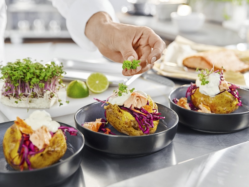 Hand eines Koches beim Anrichten mehrerer Ofenkartoffeln mit Pulled Lachs, Rotkohl und MILRAM Sour Cream