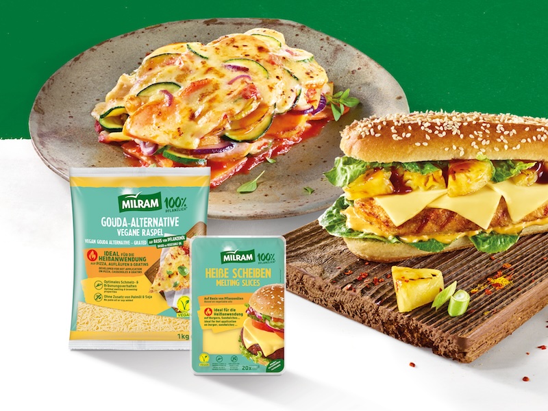 Im Vordergrund die Produktverpackungen von der MILRAM Gouda Alternative Vegane Raspel und MILRAM Heiße Scheiben; dahinter eine vegane Pizza Margherita und ein No-Chicken-Sandwich mit Salatblättern und gegrillter Ananas