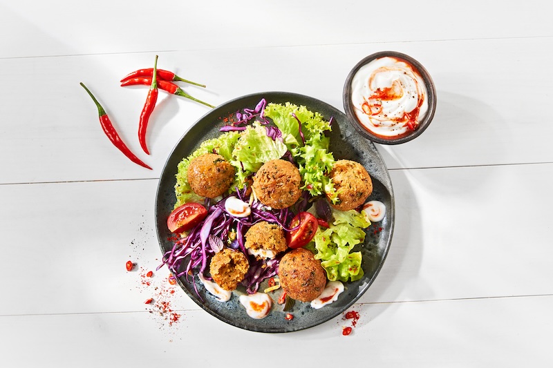 Runder Teller mit Falafelbällchen, Salat, Rotkohl und Tomate; daneben ein Schälchen mit MILRAM VeJog Joghurt-Alternative