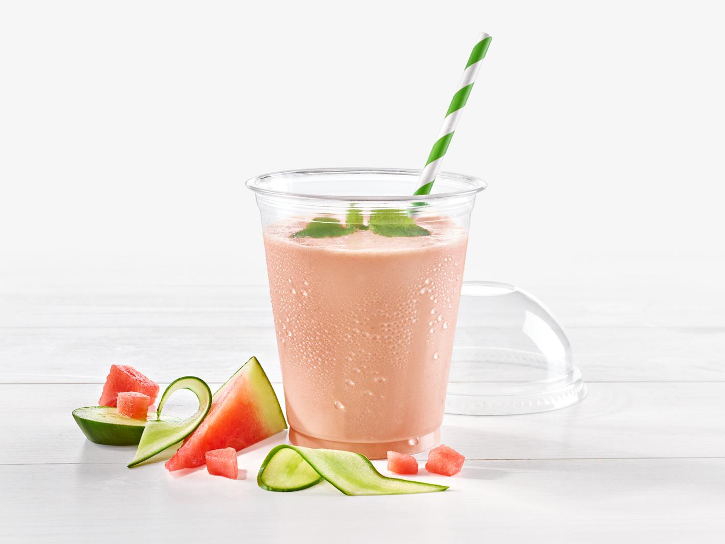 To-Go-Becher gefüllt mit Gurken-Melonen-Smoothie garniert mit einem Pfefferminzblatt; davor Gurken- und Melonenstücke