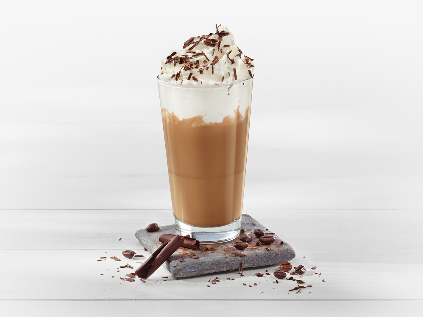 Latte Macchiato im Glas garniert mit einem Häubchen aus MILRAM Crème Brûlée, Schokoladenraspeln und Kaffeebohnen